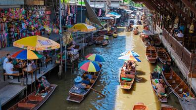 Links und rechts von einem Fluß in Thailand sitzen Menschen. Auf dem Wasser fahren Holzboote, die Menschen und Lebensmittel transportieren.