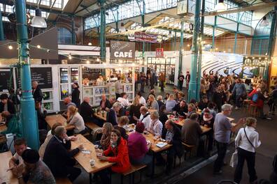 In einer altertümlichen Markthalle reihen sich Essensstände, Bierbänke und Menschen aneinander.